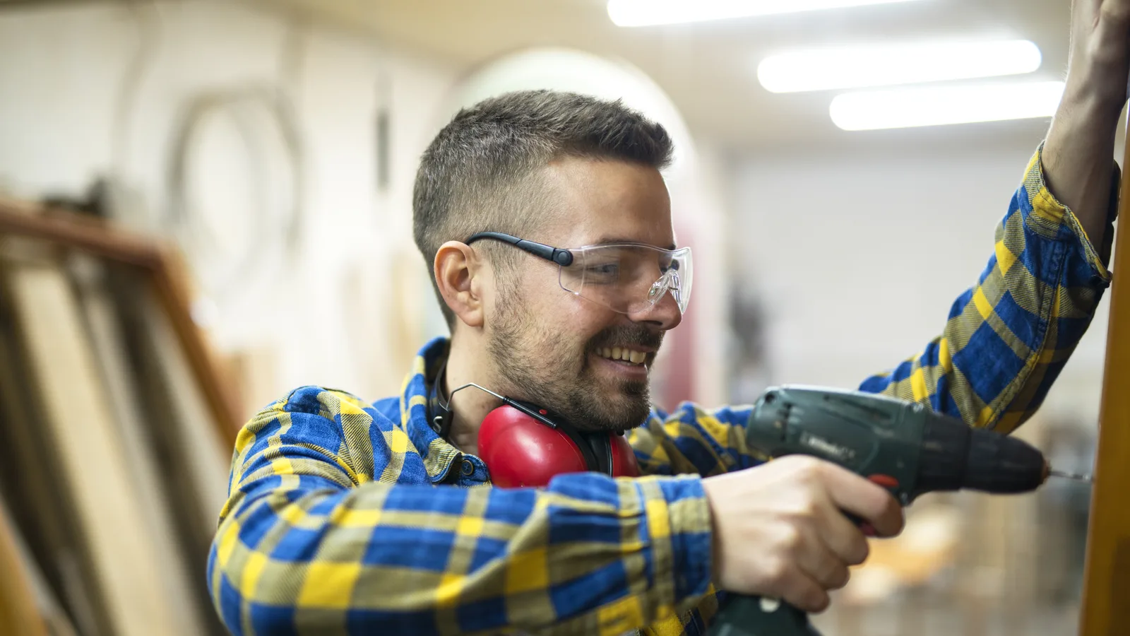 Un menuisier porte des lunettes de protection transparentes et un casque antibruit autour du cou, perceuse en main dans son atelier